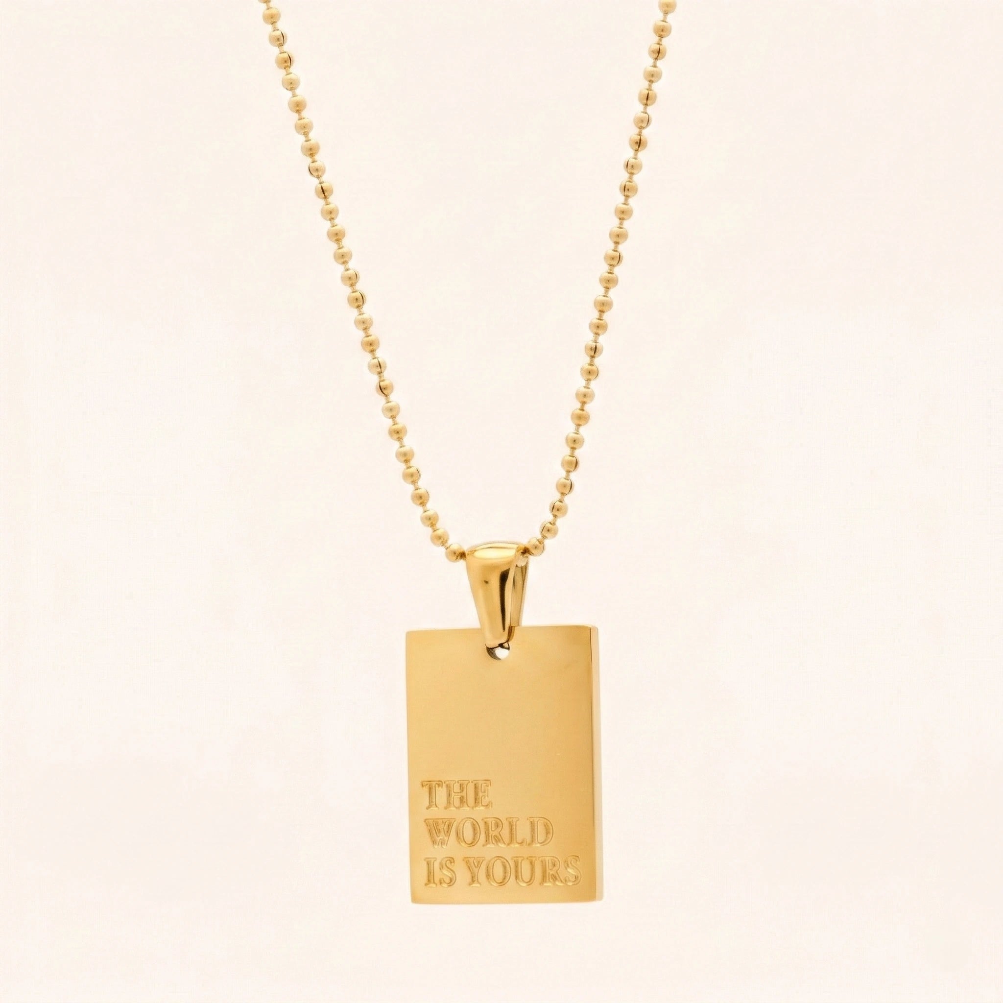 Gold necklace with a pendant that says 'The World is Yours' on a beige background